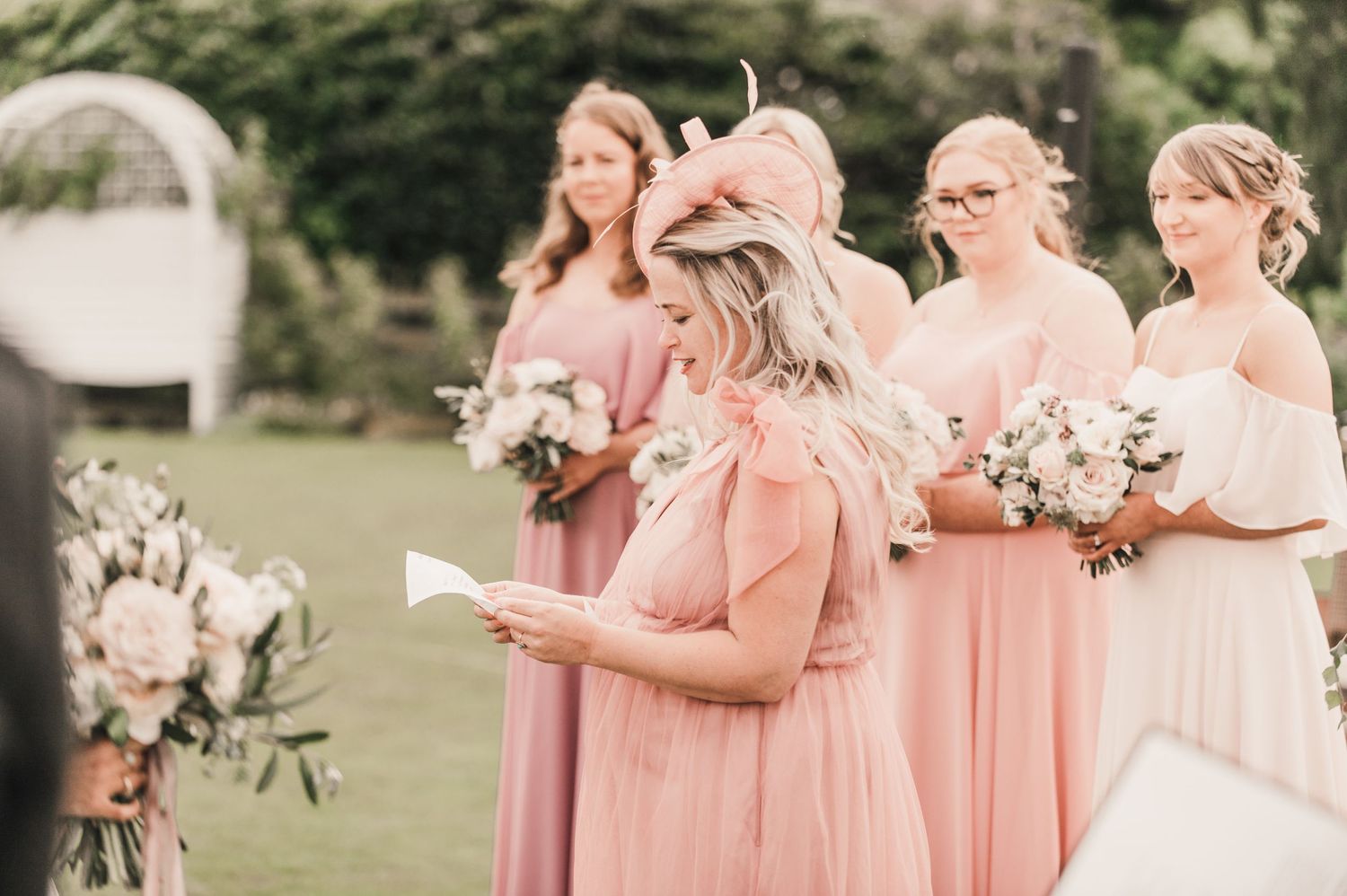 Bridesmaids in pink dresses hold bouquets during an outdoor wedding ceremony.