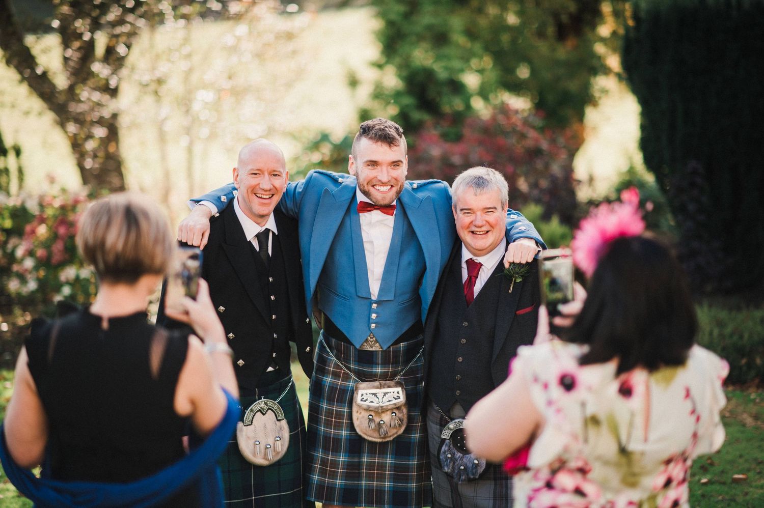 Three men in traditional Scottish kilts and formal attire smile and chat during an outdoor wedding celebration.