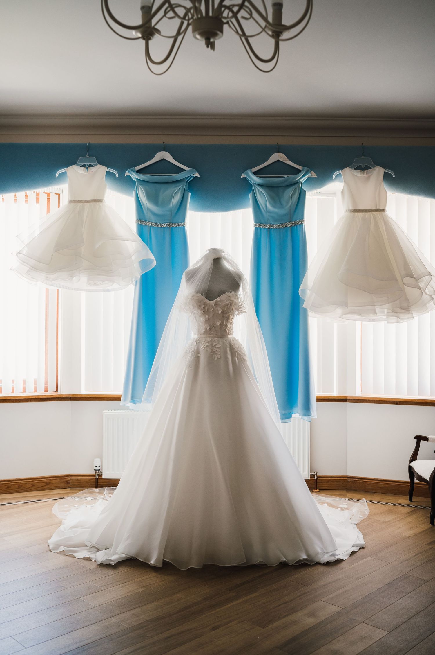 Wedding dresses and blue bridesmaid dresses hanging from a chandelier in an elegant room.