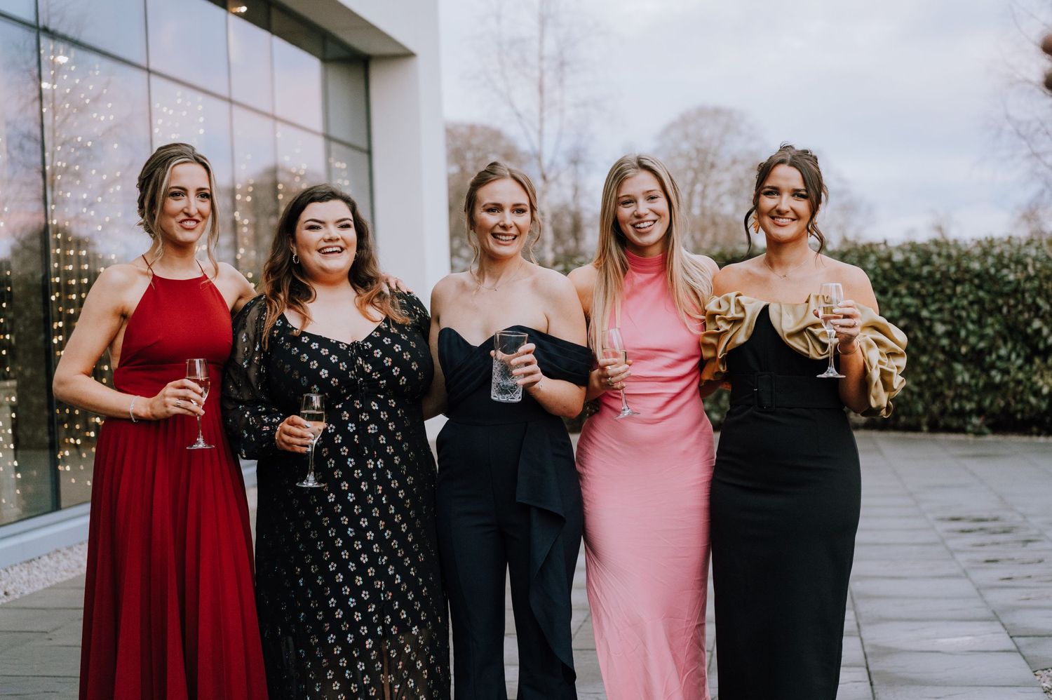 Five friends in elegant evening gowns pose together for a photo outdoors.