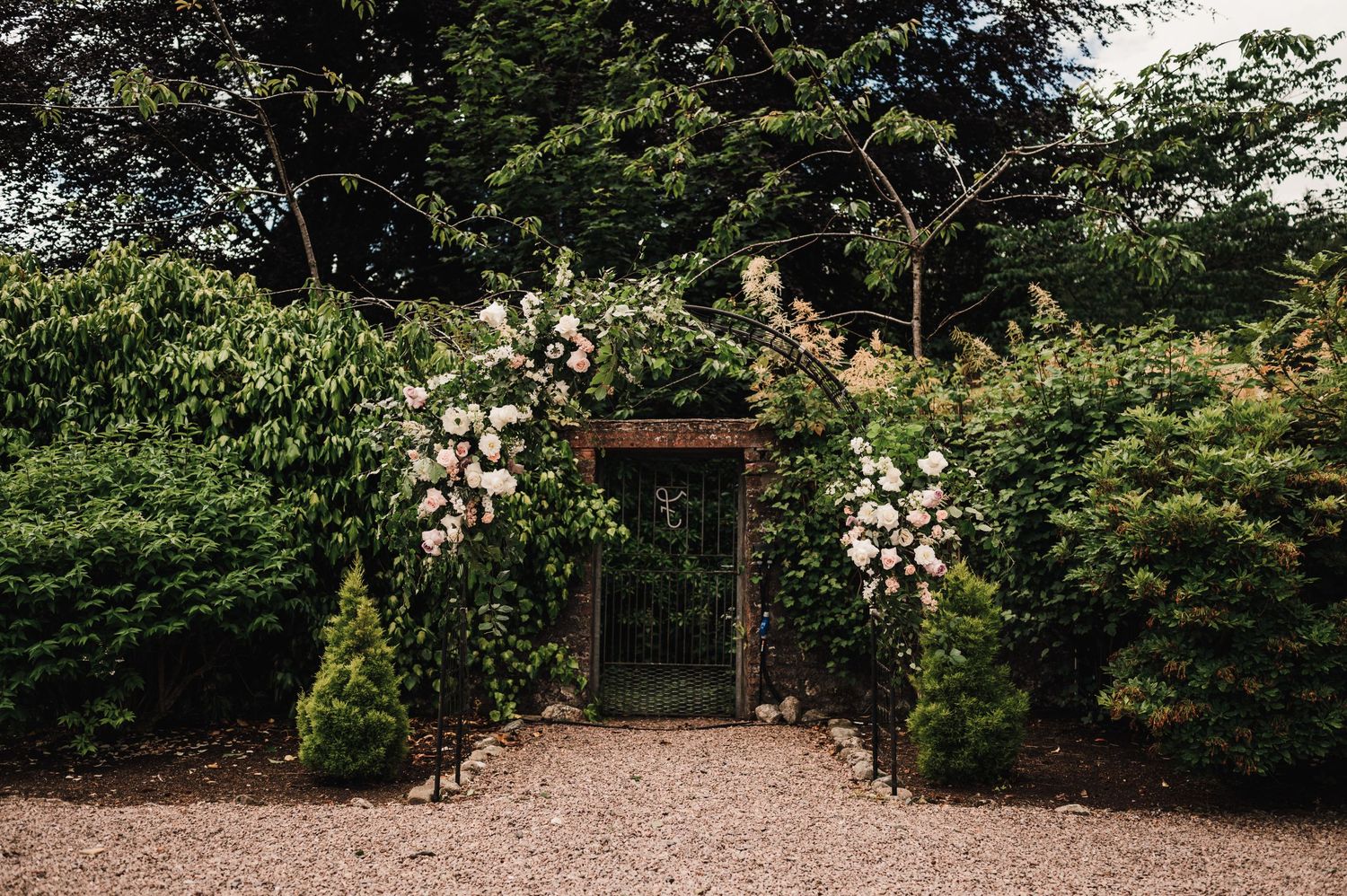 Wedding ceremony site with floral arch and gravel path surrounded by lush greenery.
