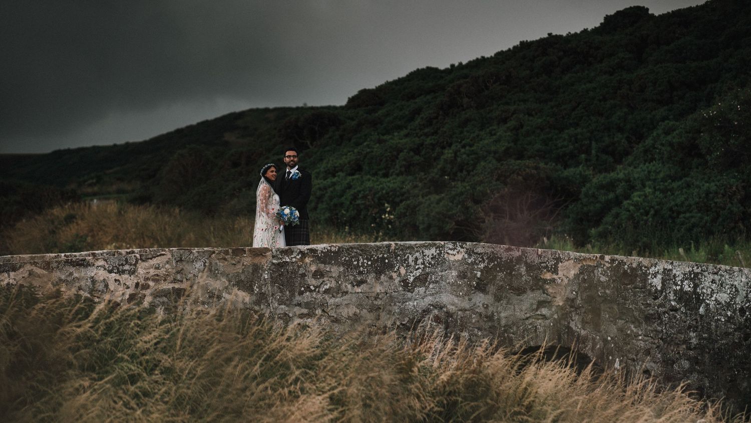 A wedding couple stands atop a stone wall against a dramatic mountainous backdrop.