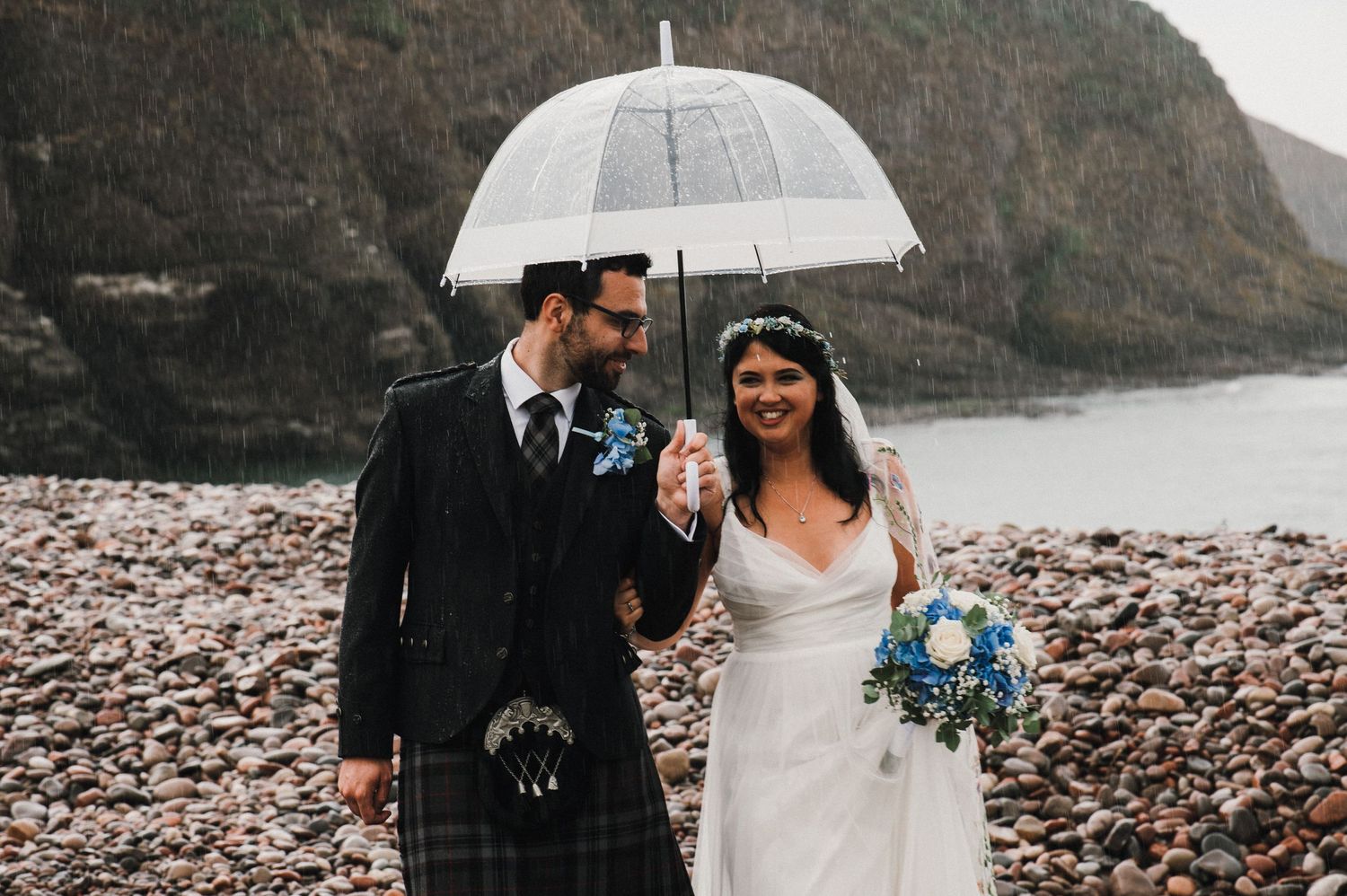 A couple in wedding attire shares an umbrella on a rocky beach, with the groom wearing a traditional kilt.