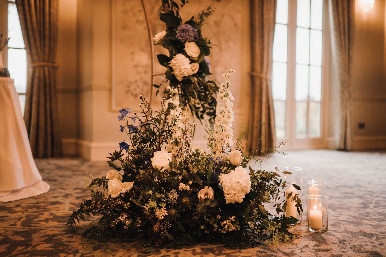 Large floral arrangement with roses and greenery against elegant wallpapered room with windows.