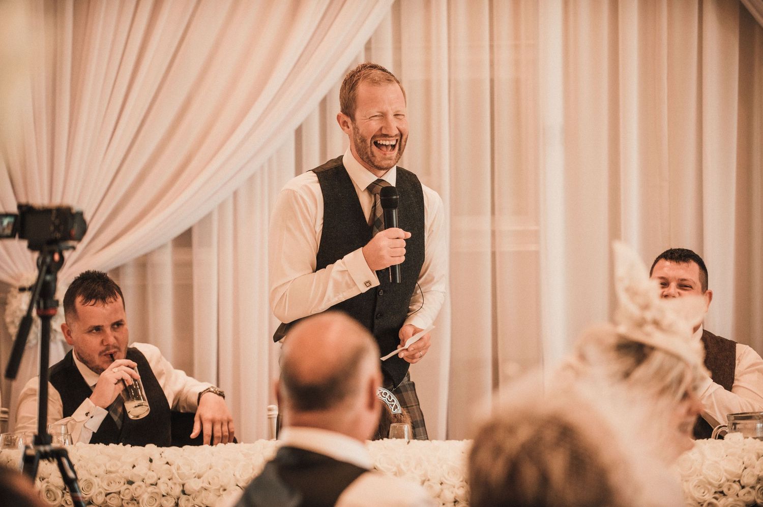 Standing speaker delivers entertaining toast causing laughter at wedding reception.