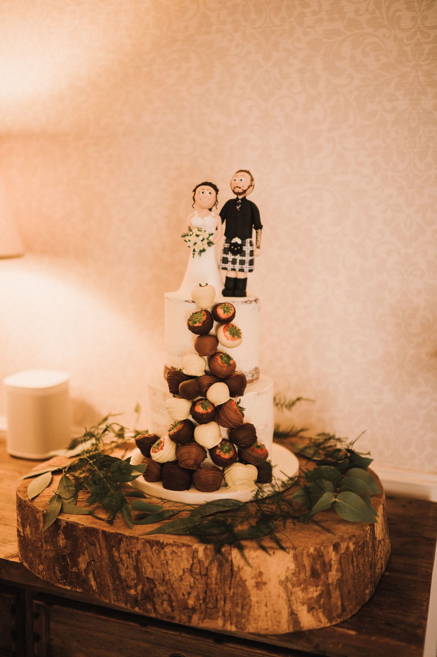 Rustic wedding cake display with custom toppers on wood slice base decorated with greenery.