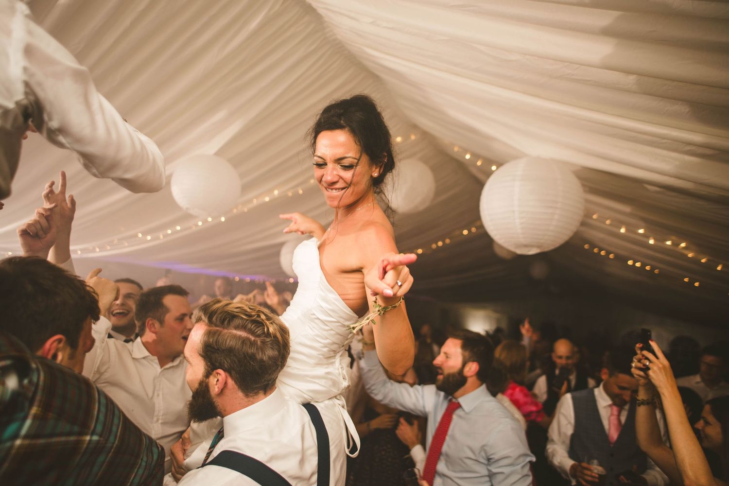 A celebratory moment at a wedding reception as guests dance under a white tent with fairy lights.