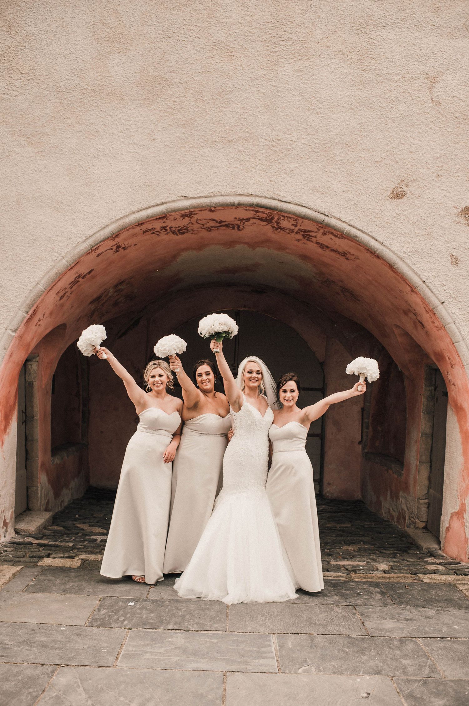 Bridal party celebrates with raised bouquets under a curved stone archway.