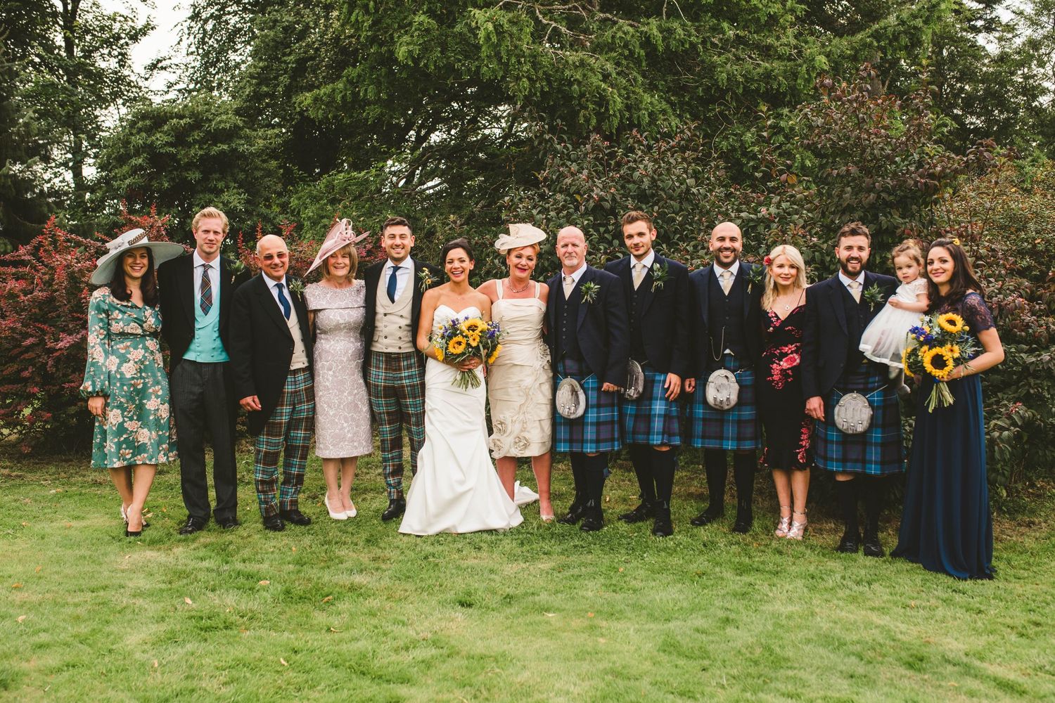 Large wedding party group photo with Scottish kilts and formal wear against garden backdrop.