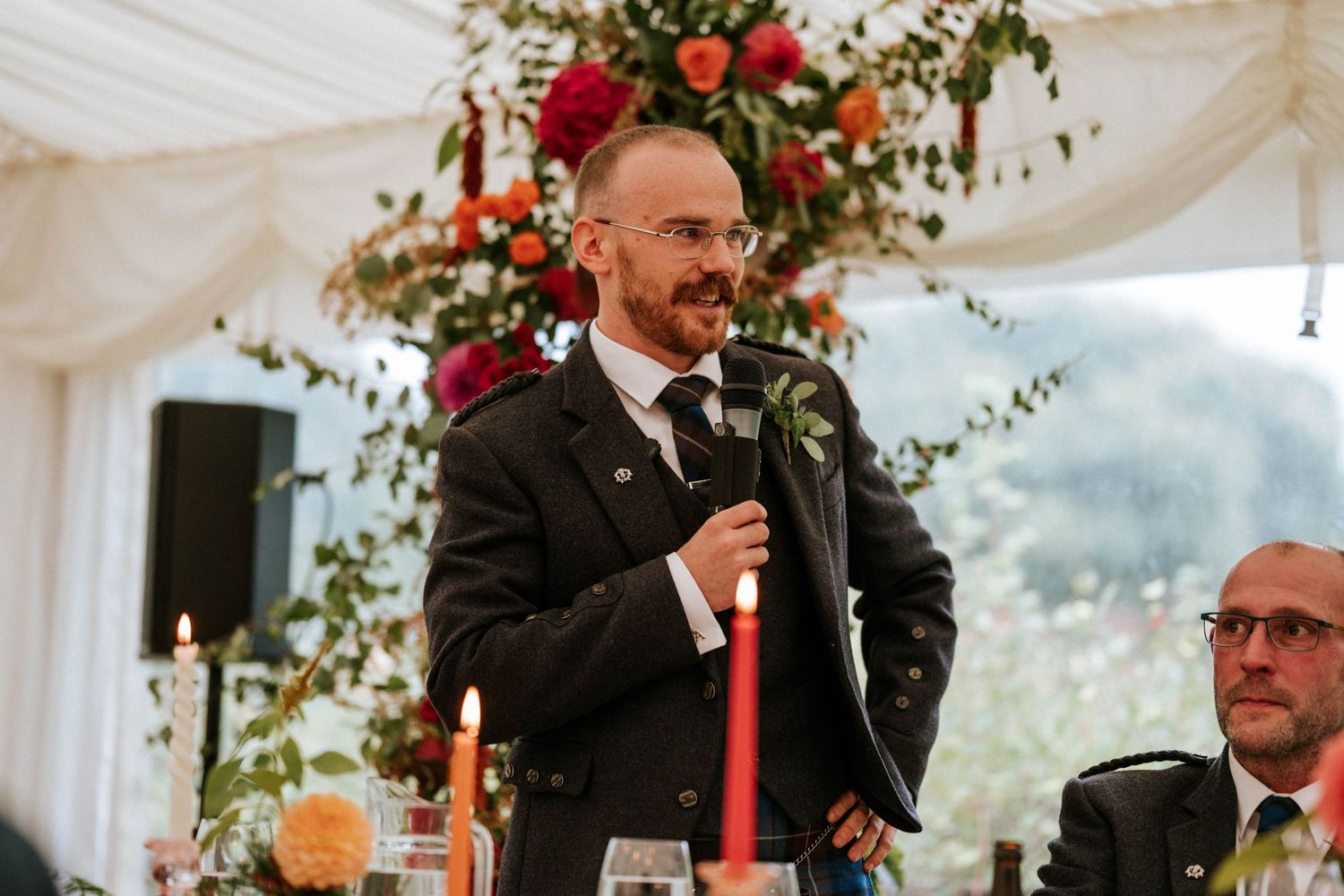 Wedding reception speeches and toasts under white draped tent with red floral arrangements.