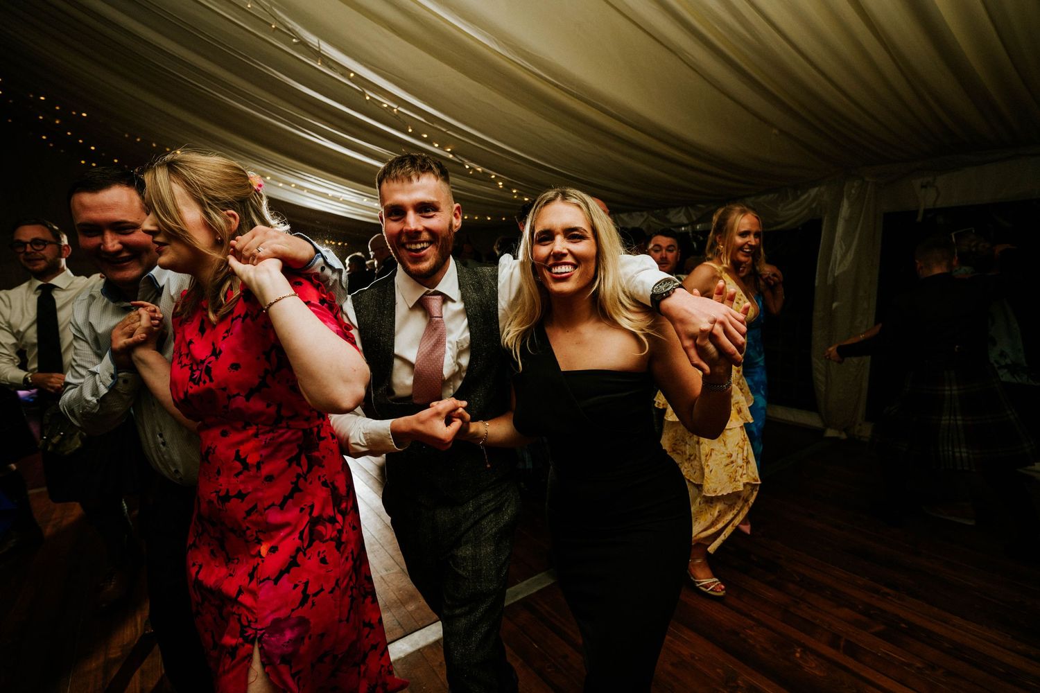 Lively dance floor scene at wedding reception with guests enjoying themselves under white tent.