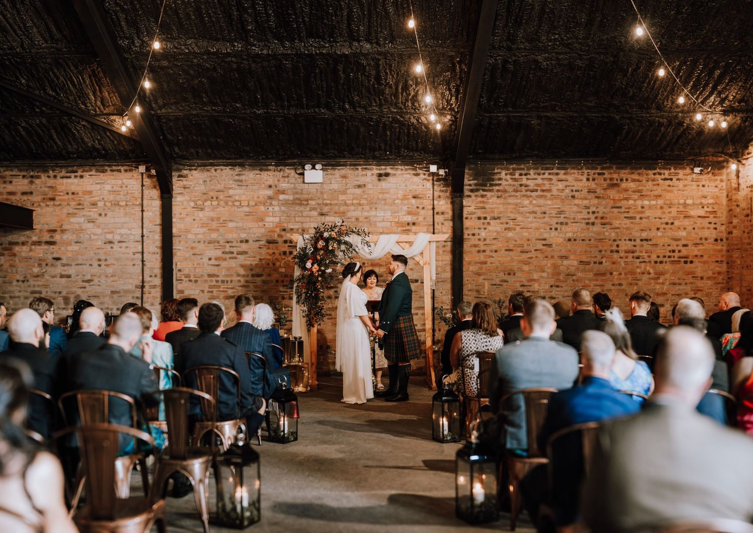 Wedding ceremony in industrial venue with string lights and exposed brick walls.