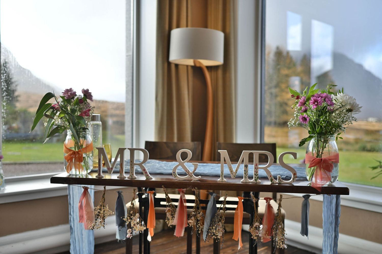 Wedding decor featuring 'MR & MRS' sign with floral arrangements on display table by window.