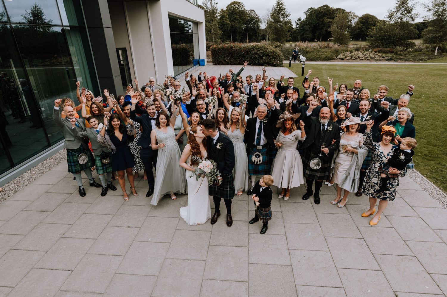 Large group wedding photo with guests cheering outside a modern building.