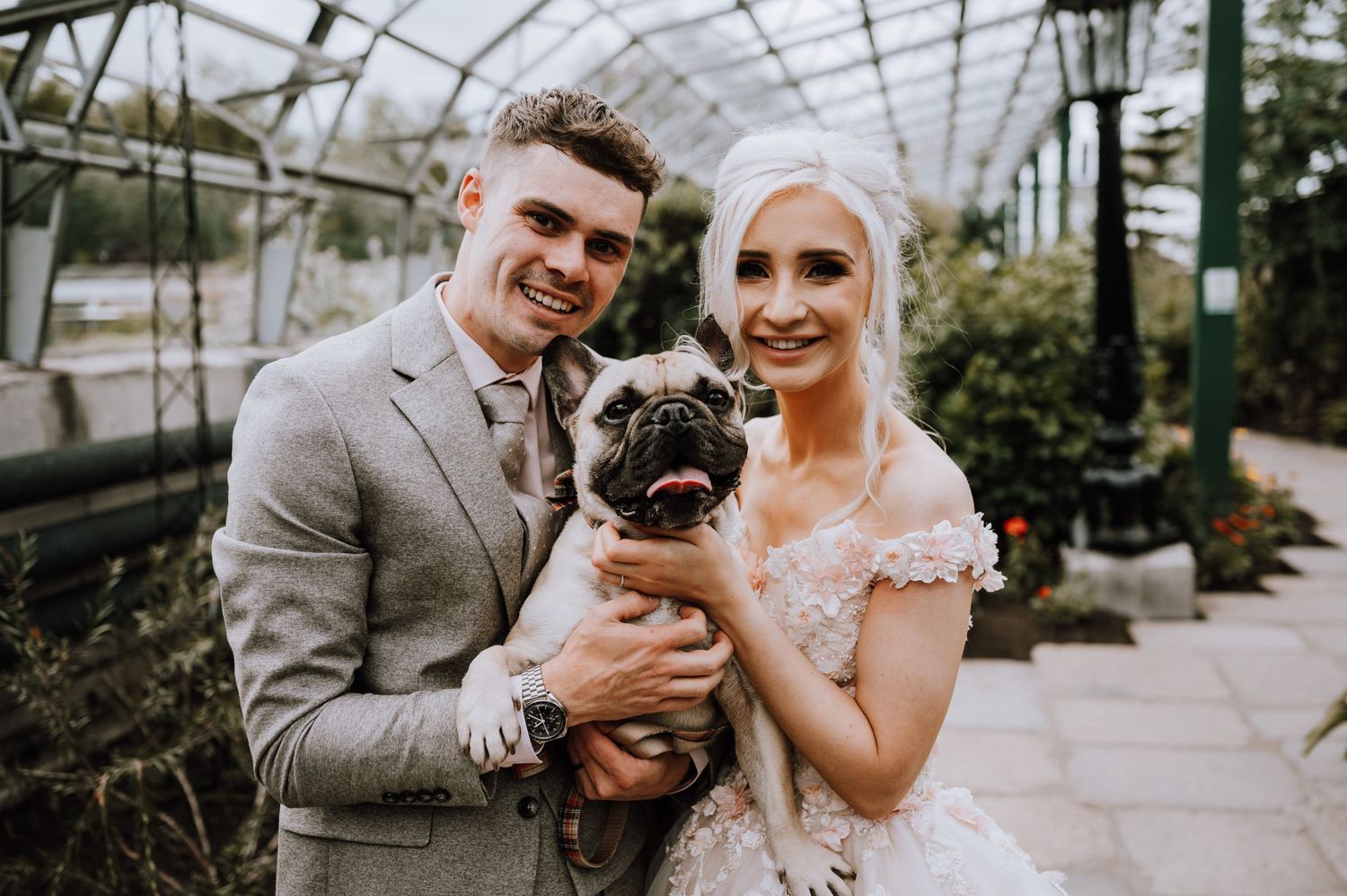 A couple in wedding attire holds their pug dog in a greenhouse setting.