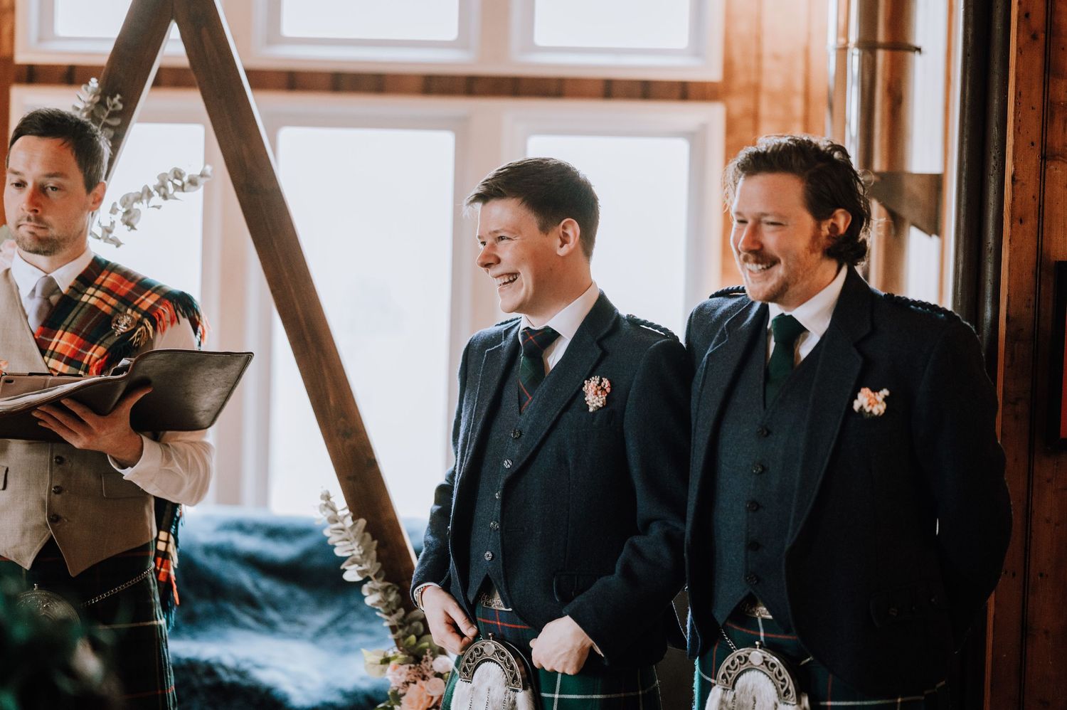 Two guests in dark suits share a laugh during a wedding ceremony in a rustic wooden venue.
