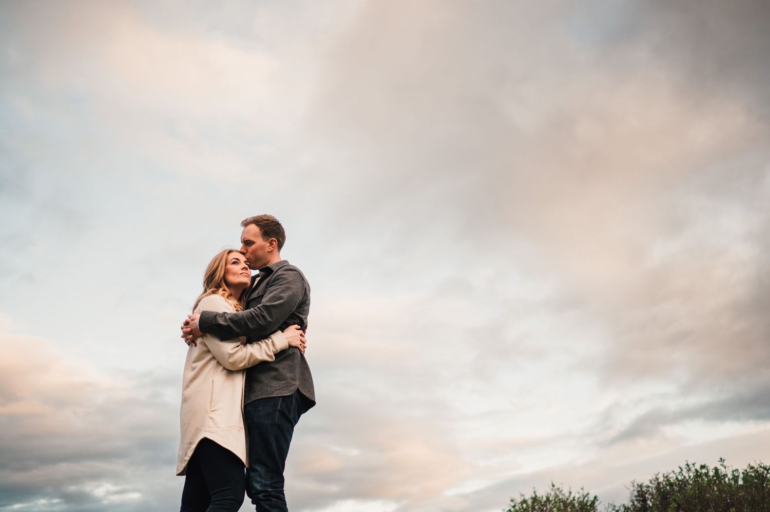 A couple shares an intimate embrace against a pink-tinged cloudy sky at dusk.