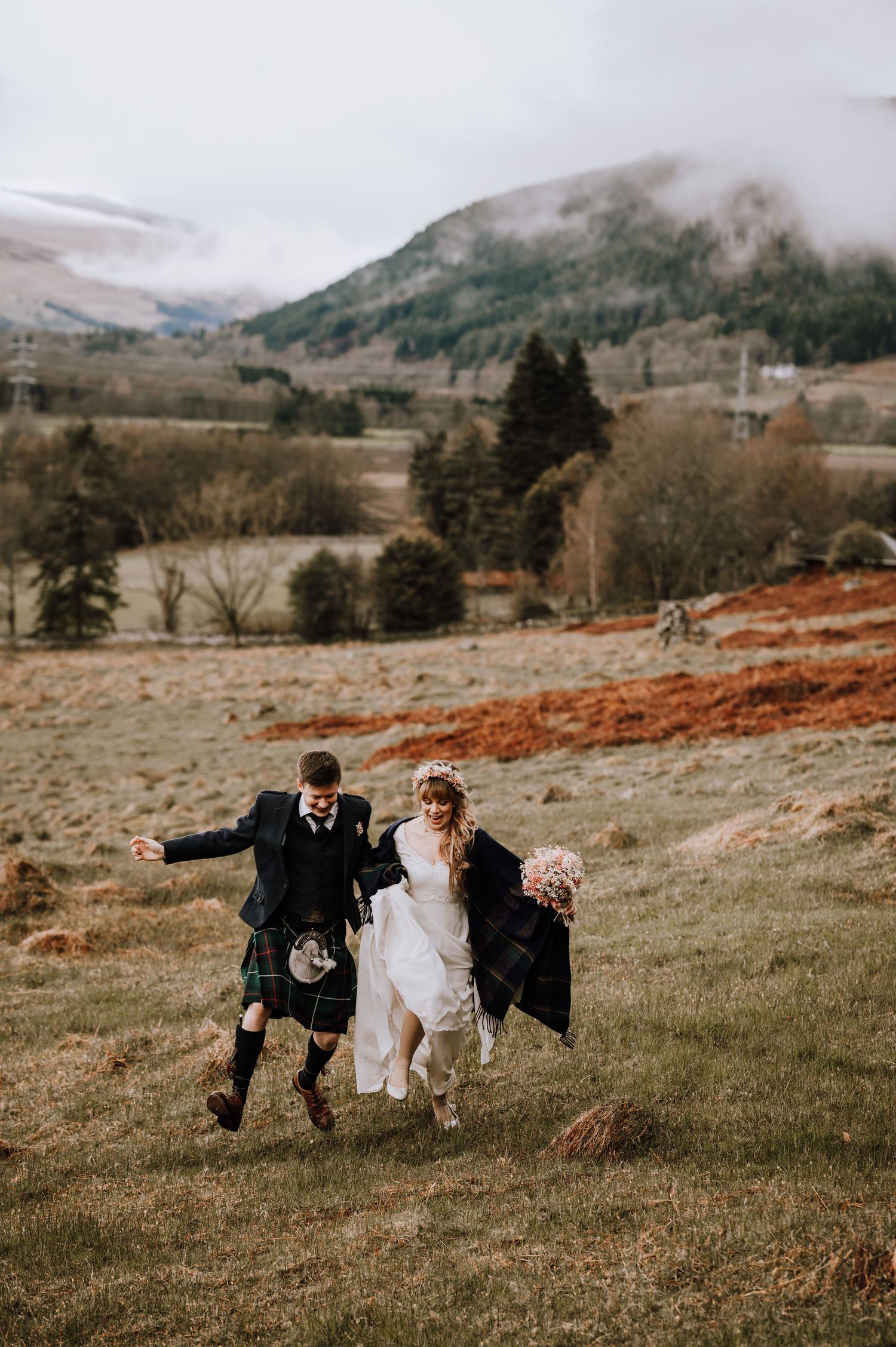 Joyful couple runs through Scottish highlands in wedding attire with fog-covered mountains behind them.
