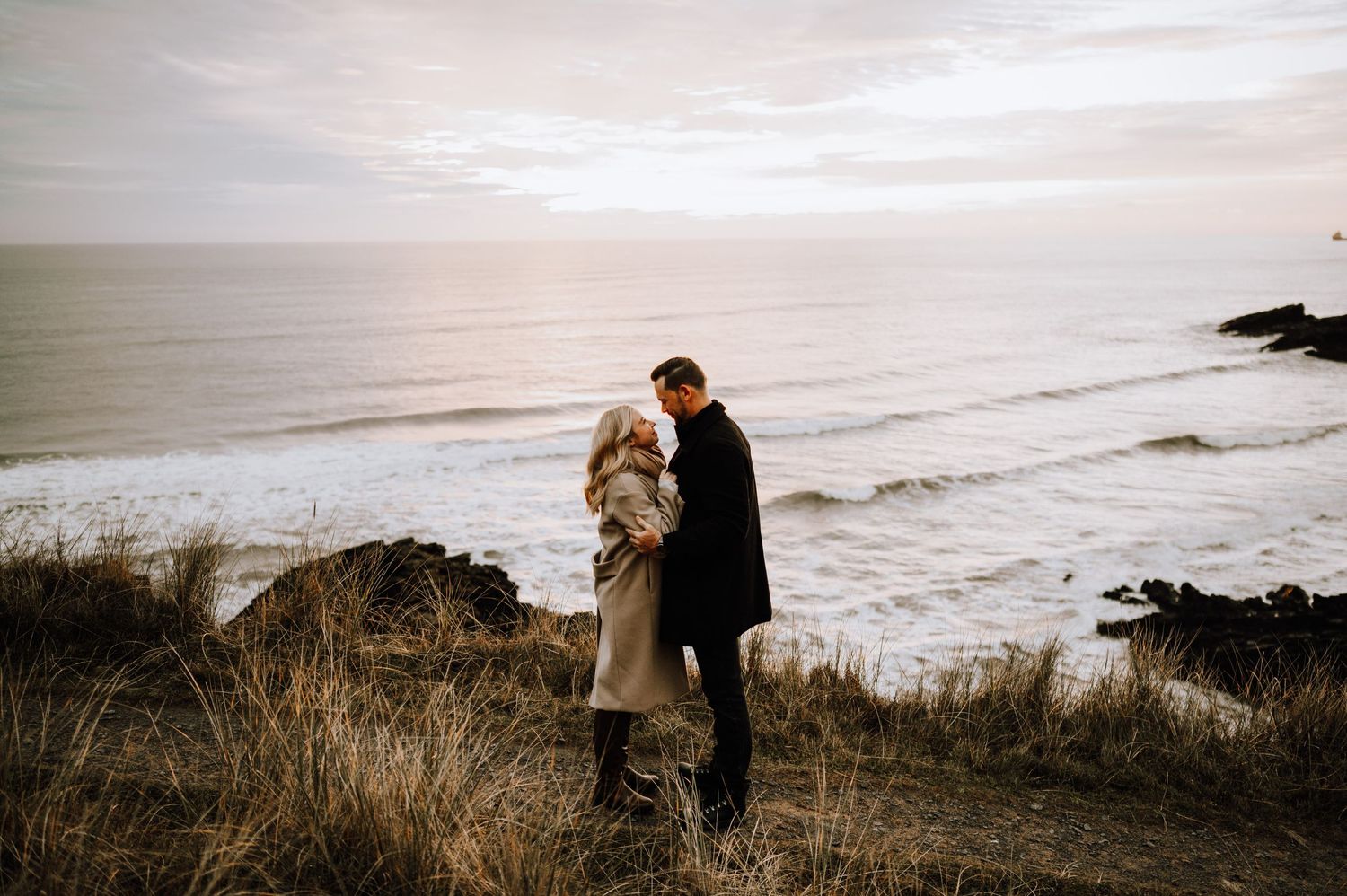 Couple embracing on coastal cliffs overlooking a dramatic ocean view at dusk.