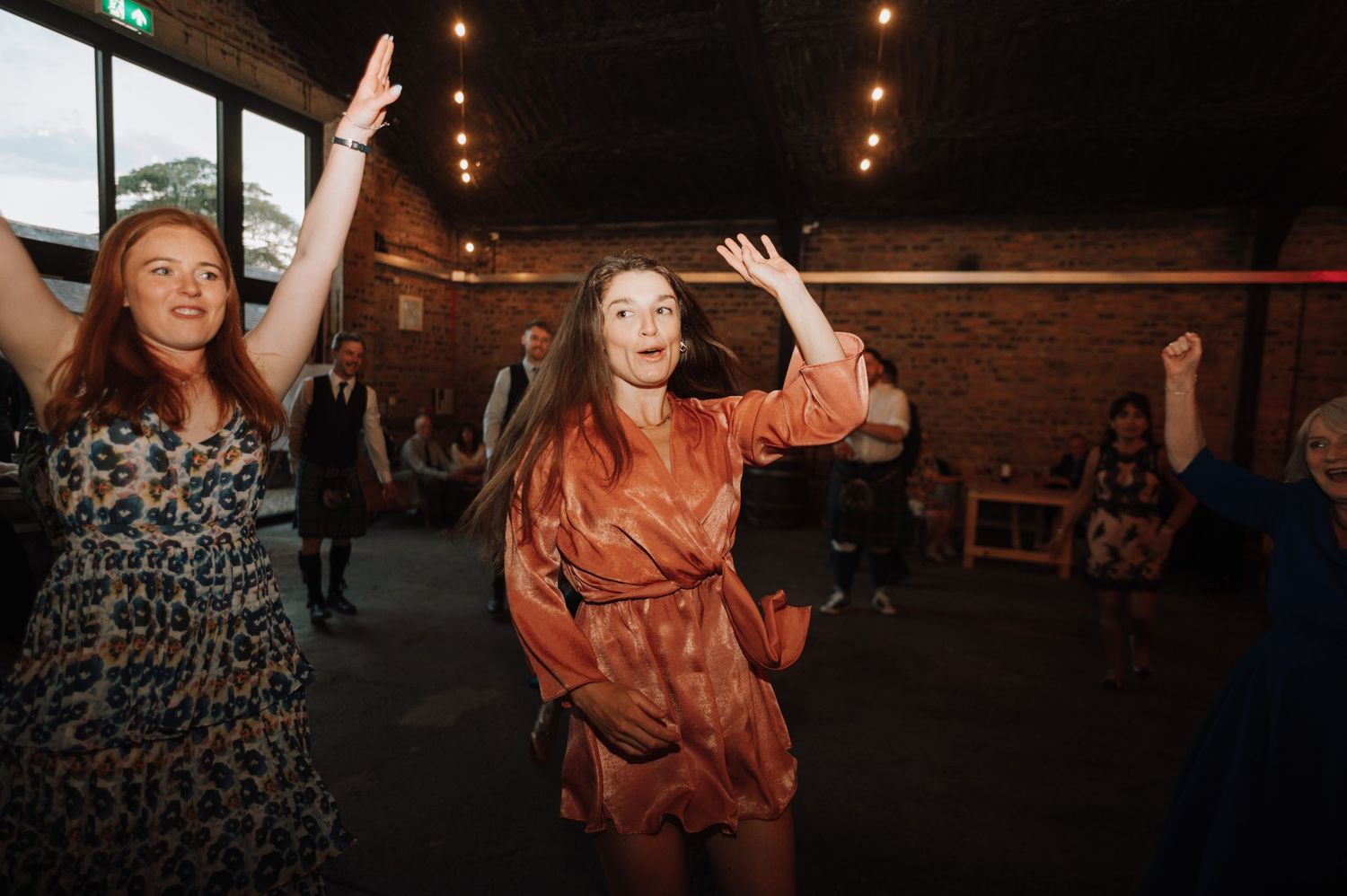 Dancing guests celebrate with raised arms at a rustic indoor reception with string lights.
