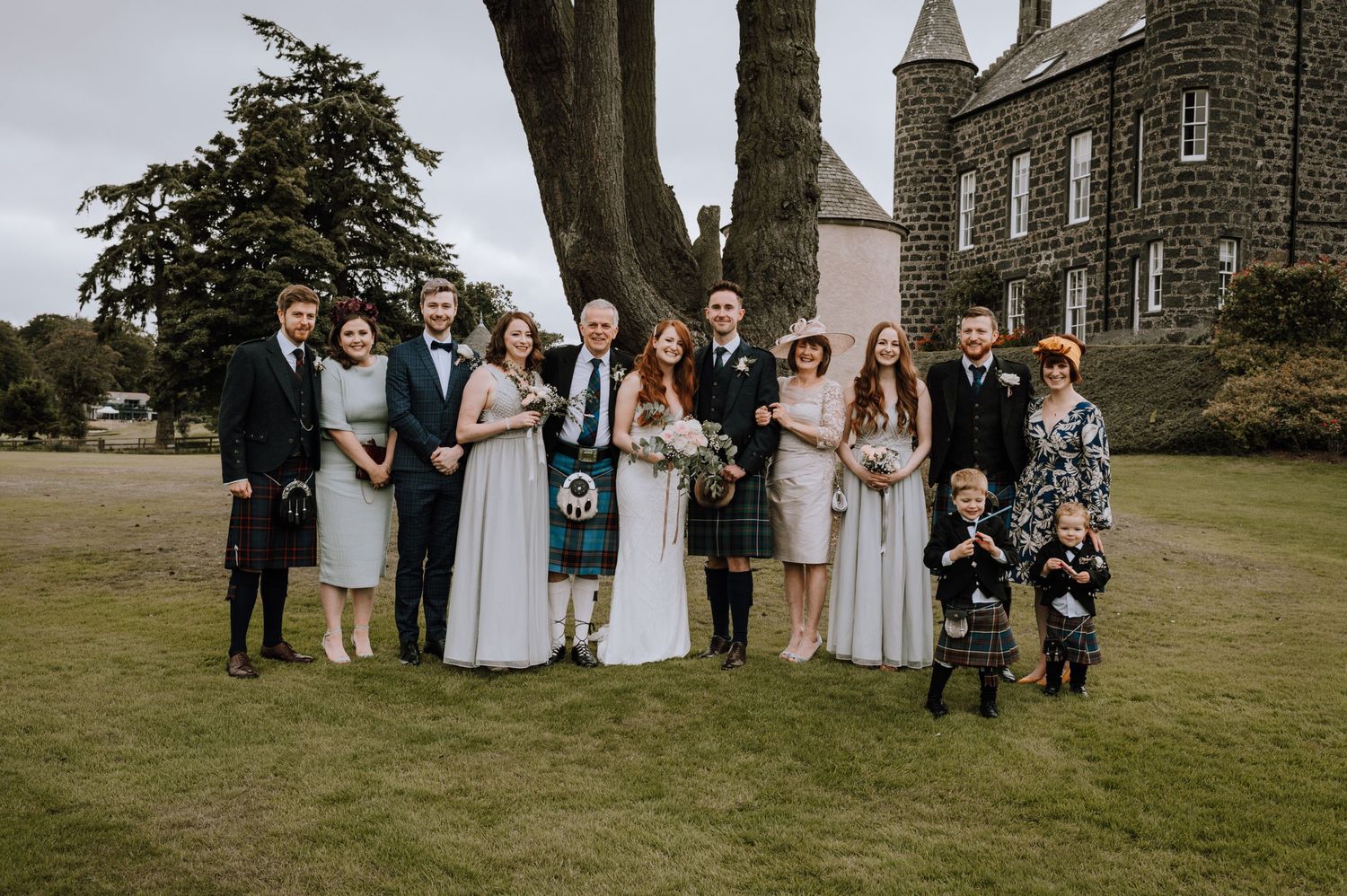 Wedding party pose for formal portrait on castle lawn wearing mix of kilts and formal dresses.