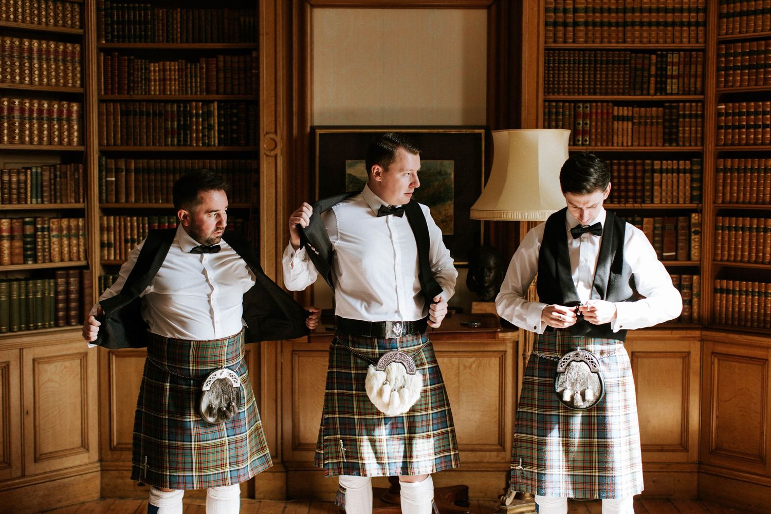 Four people in traditional Scottish kilts and formal attire pose in a grand library with wooden bookshelves.