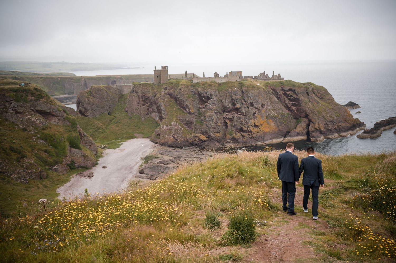 Same Sex Wedding Photography. Two figures stroll together on a grassy coastal path with rugged cliffs and castle ruins in the background.
