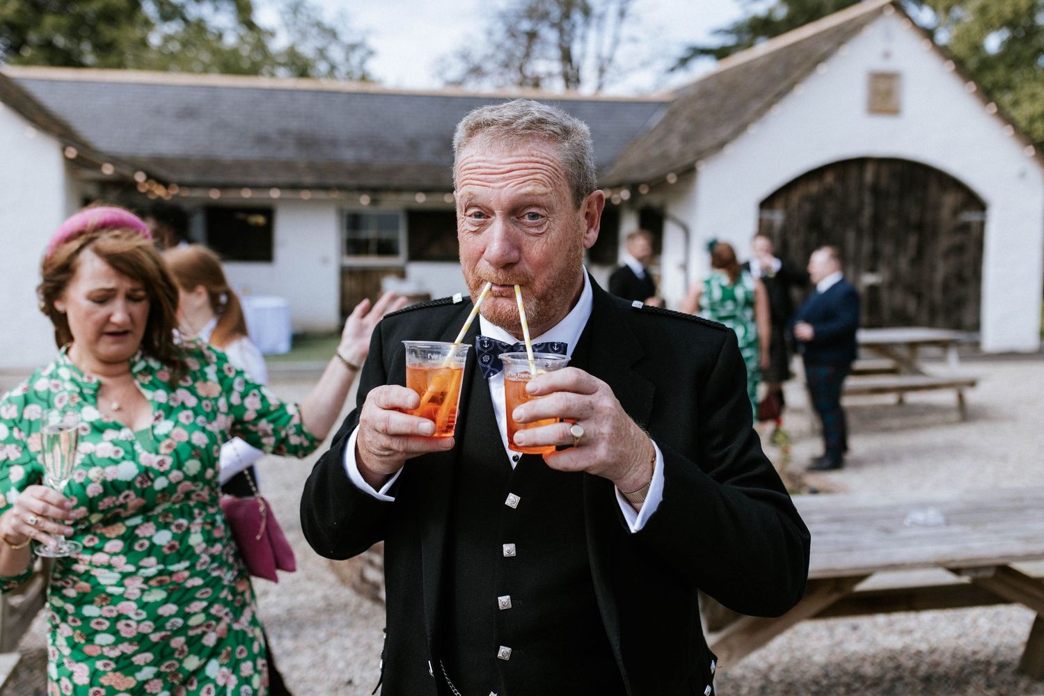 Aberdeen Wedding Photography. Guest in traditional Scottish attire enjoying two drinks at an outdoor wedding reception.