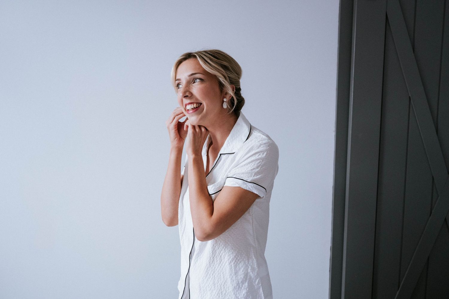 A bride in a white dress stands near a window in an emotional first look moment.