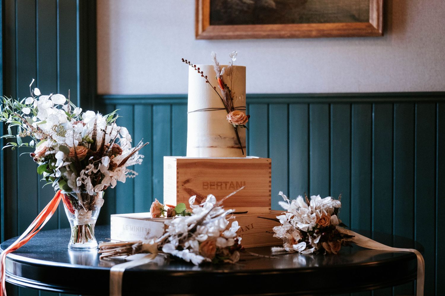 A modern three-tiered wedding cake with dried floral arrangements displayed on a dark round table against teal paneled walls.