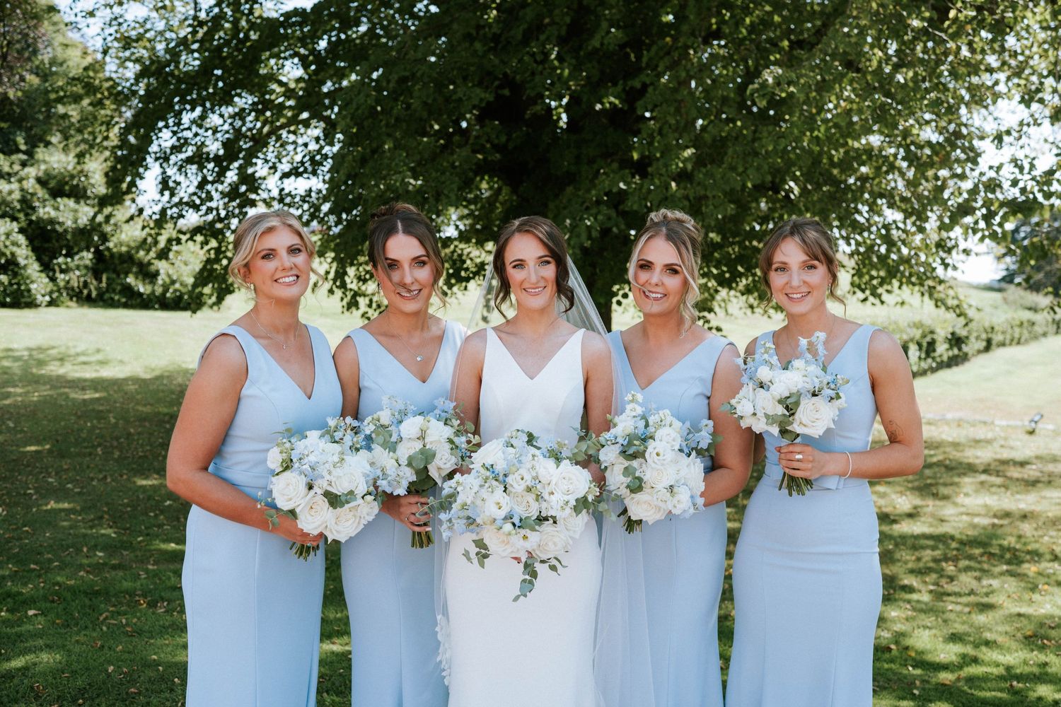 Bridesmaids in light blue dresses pose together outdoors holding white floral bouquets.