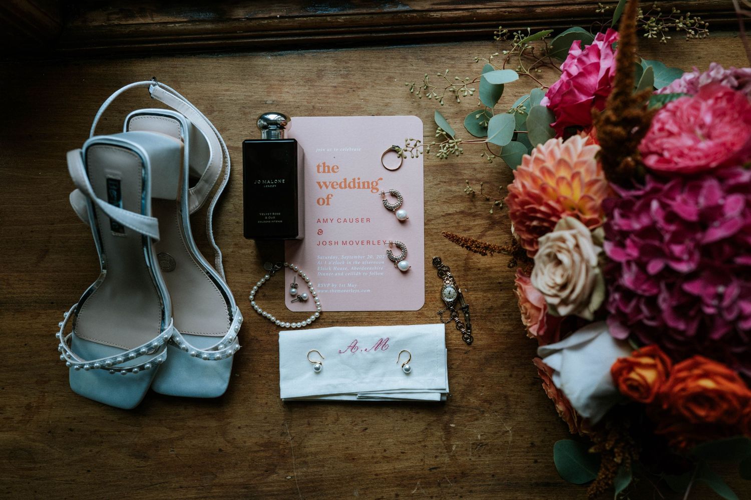 Wedding details including floral arrangements, blue shoes, and invitation cards laid out on rustic wooden surface.