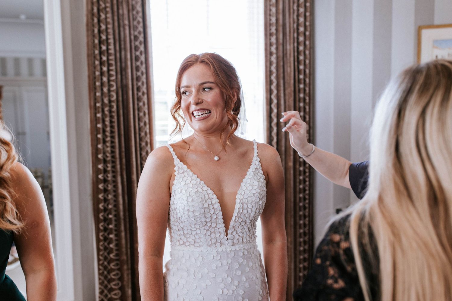 A bride in a white beaded wedding dress with plunging neckline gets ready in front of an ornate mirror.