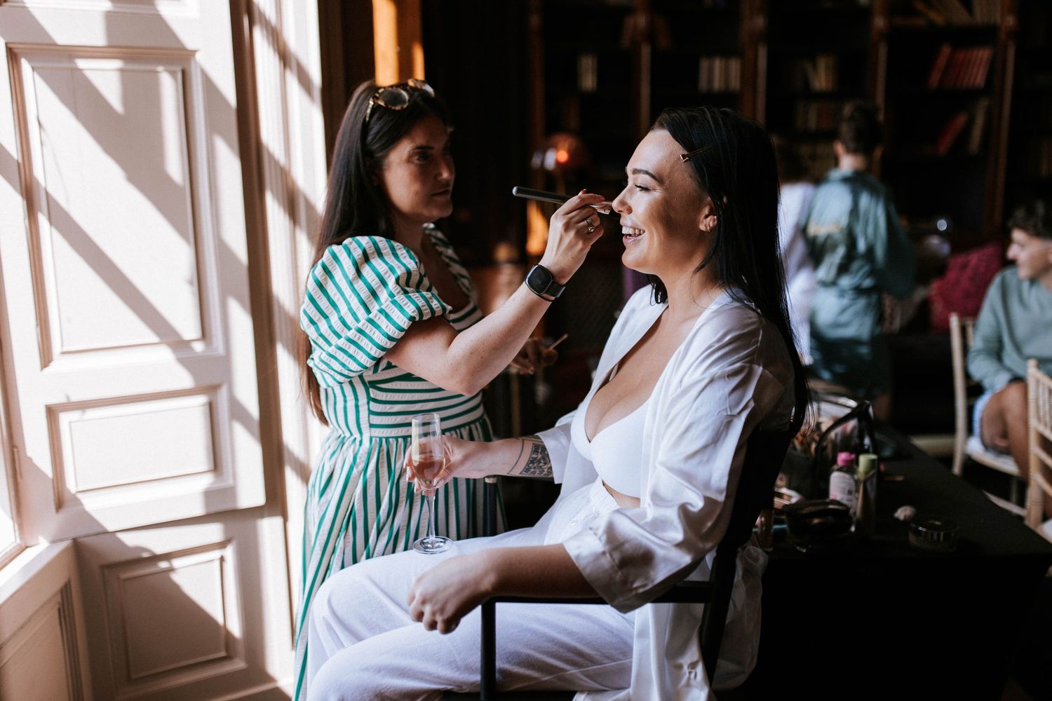 Makeup artist applying cosmetics on a client in a sunlit room during wedding preparations.