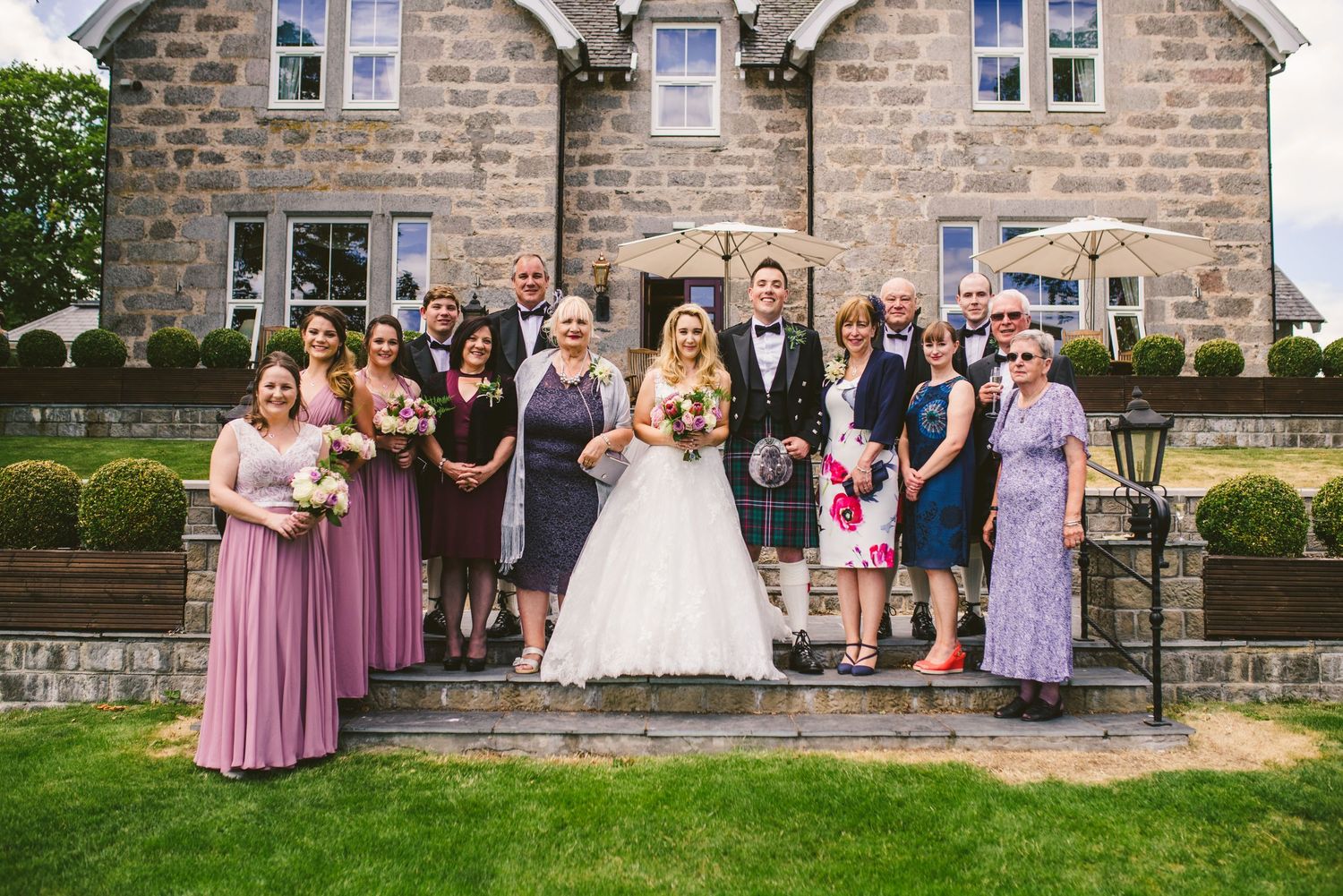 Wedding group photo outside stone country house with manicured lawn.