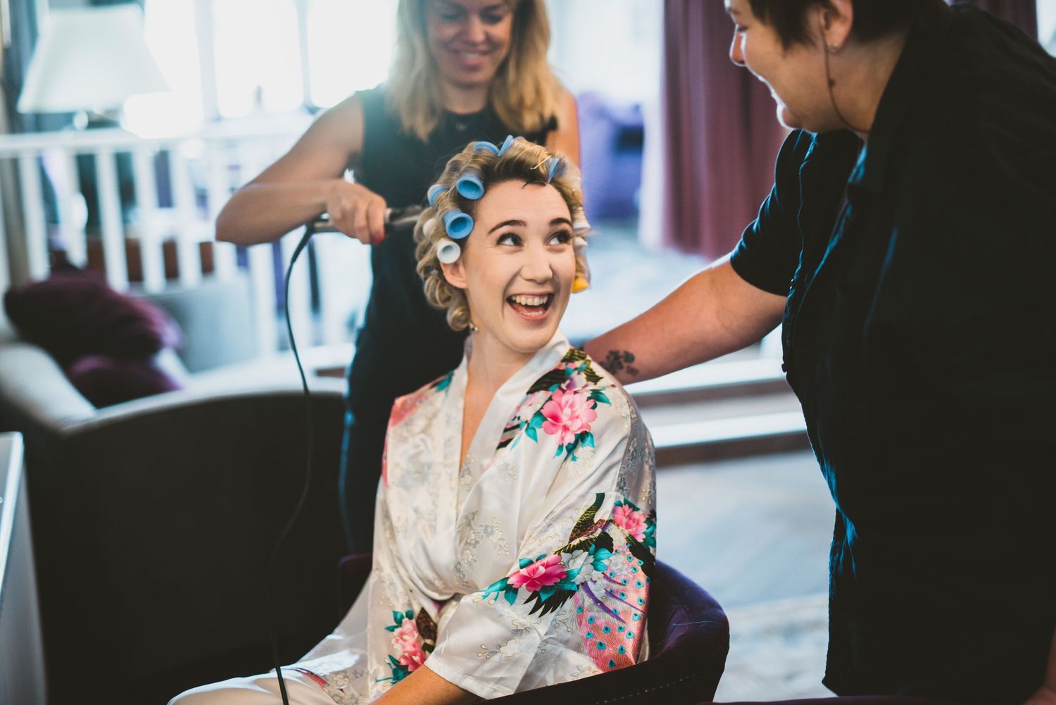 A person wearing a floral robe sits in a chair smiling while getting their hair styled at a salon.