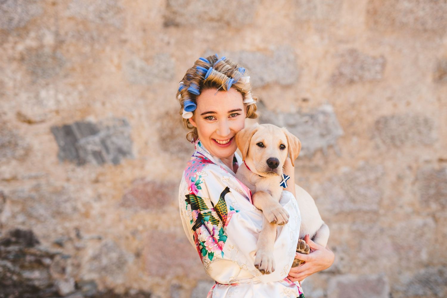 Woman in floral dress poses with yellow Labrador puppy against stone wall backdrop.