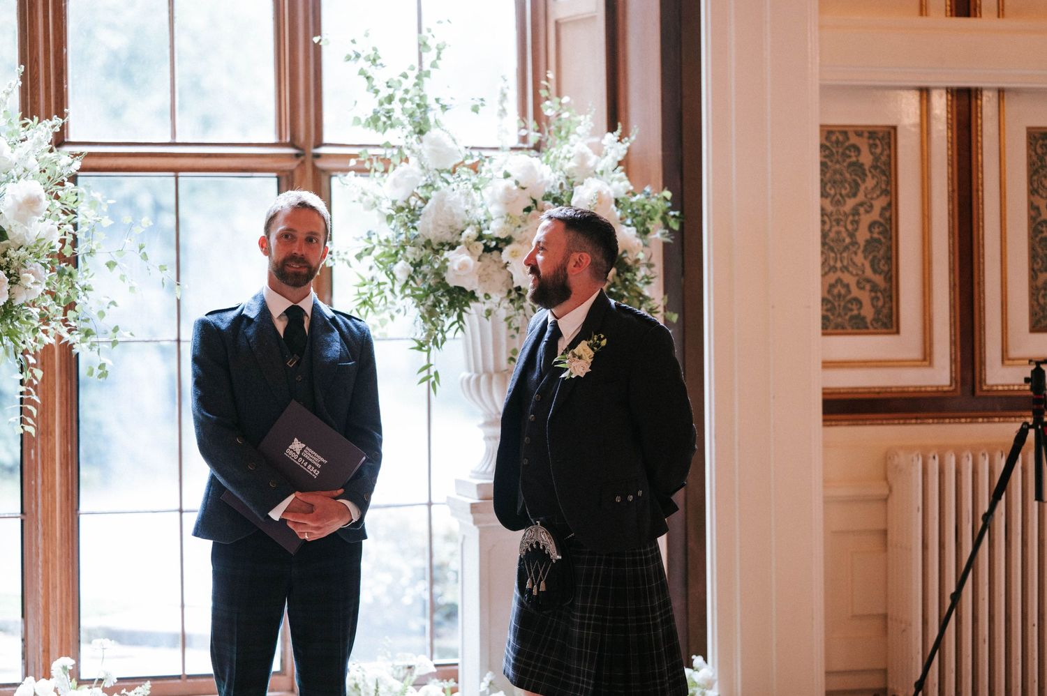 Wedding guests in formal attire standing in an elegant venue with large windows.