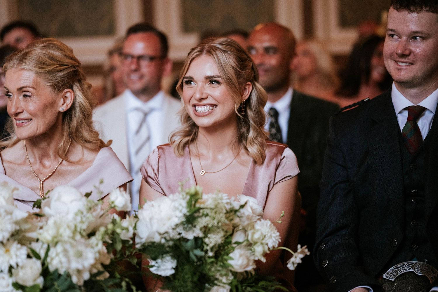Wedding party members smile and laugh during an elegant indoor reception with floral arrangements.