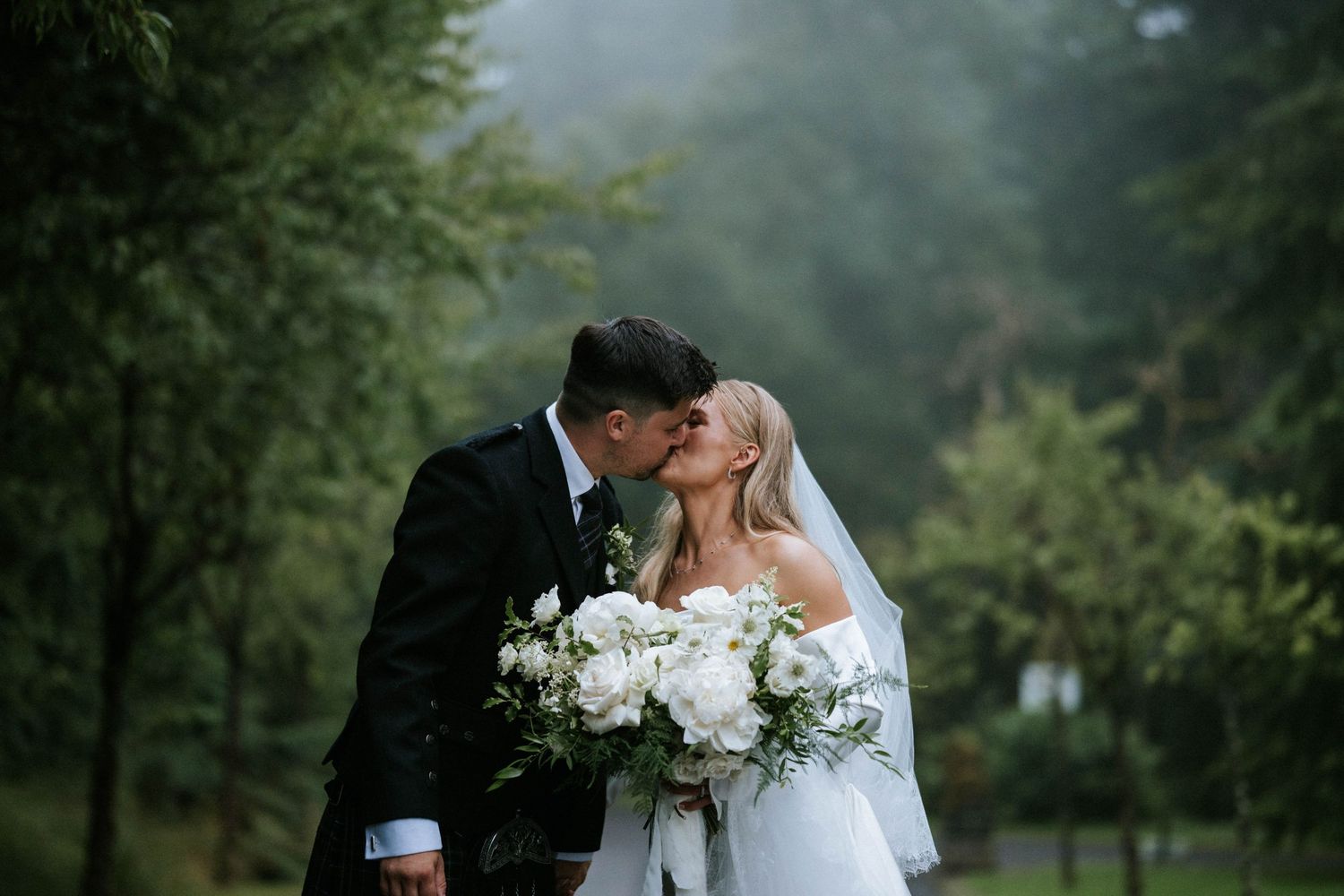 Newlyweds share intimate moments on a foggy mountain path with white floral bouquet.