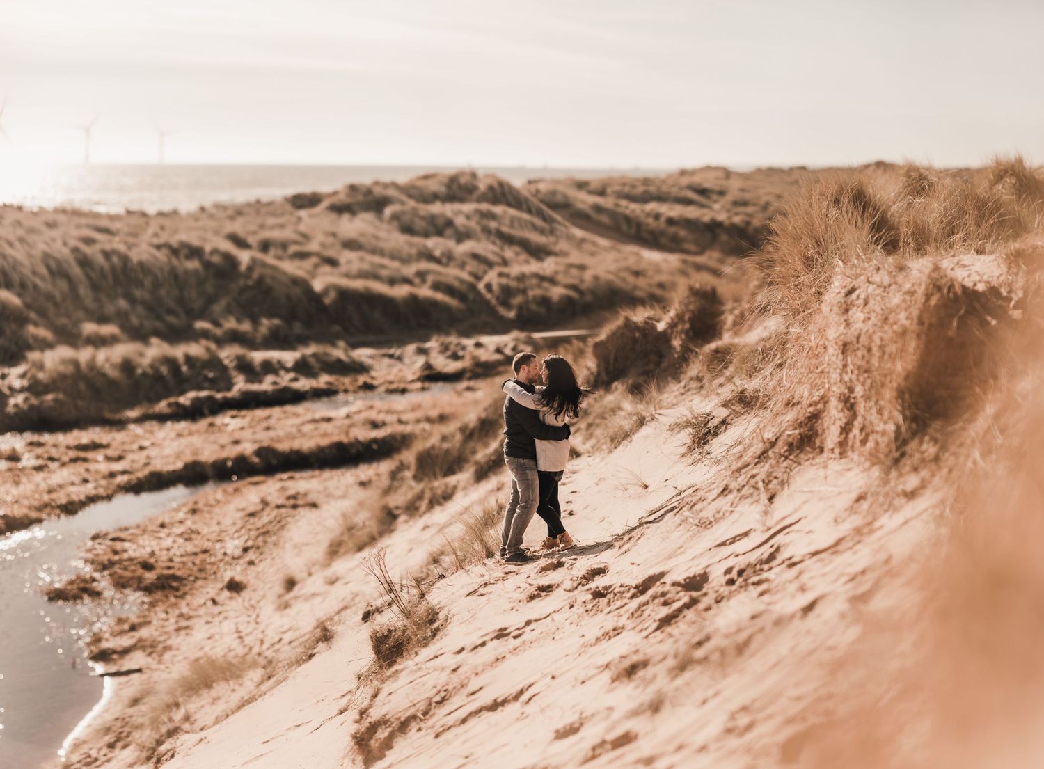 A couple in wedding attire stands on a sandy hillside overlooking a scenic landscape at sunset.