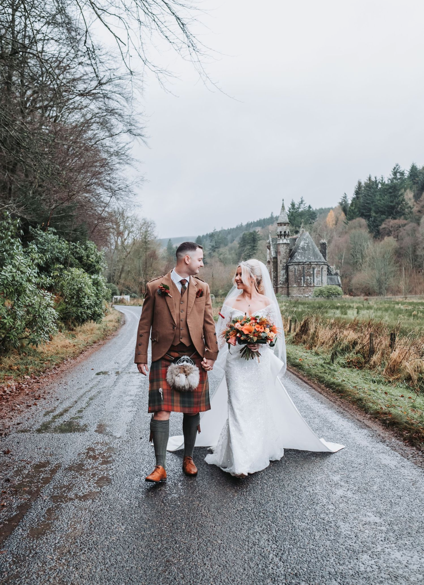 A couple walks along a misty country road in Scotland wearing traditional wedding attire and Scottish Highland dress.