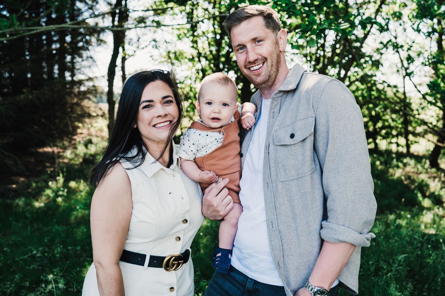 A family wearing light colored clothing poses together outdoors in a natural setting with green foliage in the background.