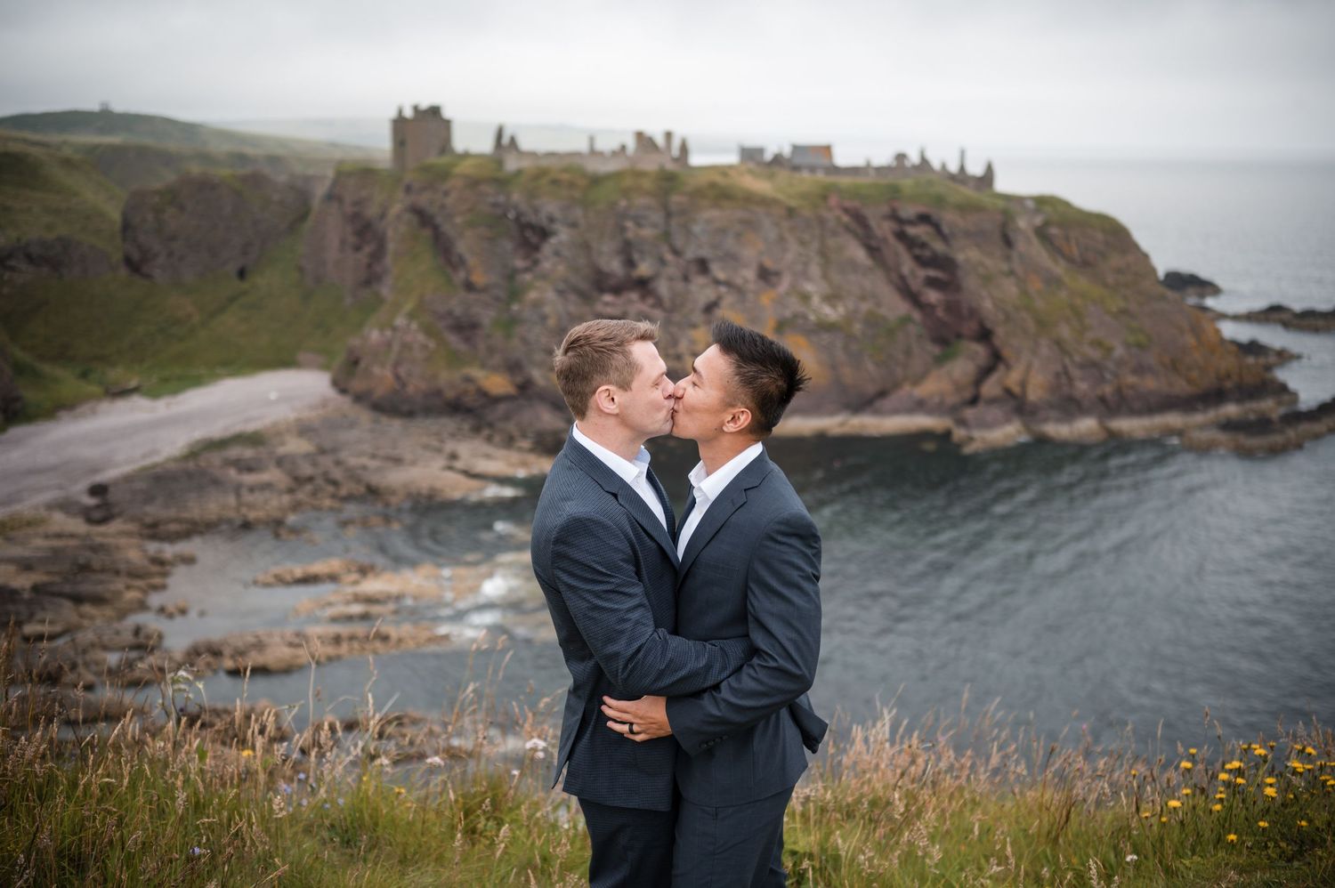 Same Sex Wedding PhotoA romantic couple shares a tender moment on a grassy cliff overlooking a dramatic coastal landscape with castle ruins.