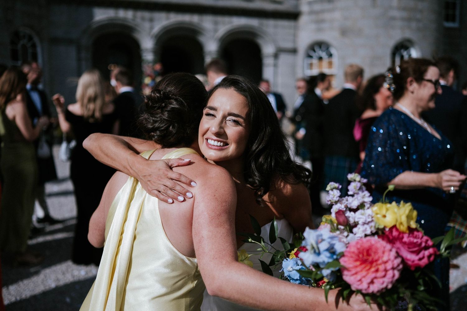 Candid moment of wedding guests hugging with vibrant floral bouquet.