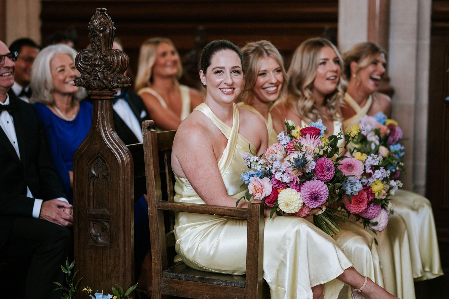 Wedding party members in pale yellow dresses seated in wooden church pews.