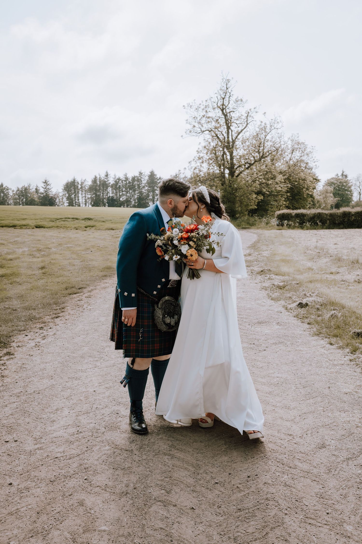 Wedding couple in traditional Scottish attire share an intimate moment on a country path.