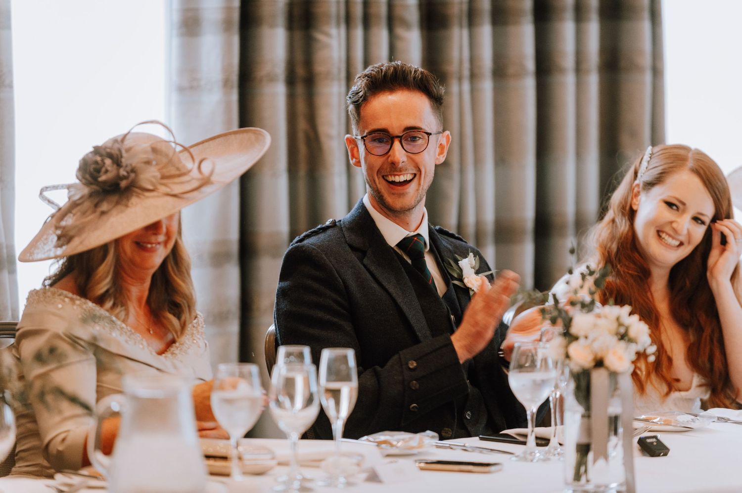 Wedding guests enjoy speeches at an elegant reception with floral decor and wine glasses.