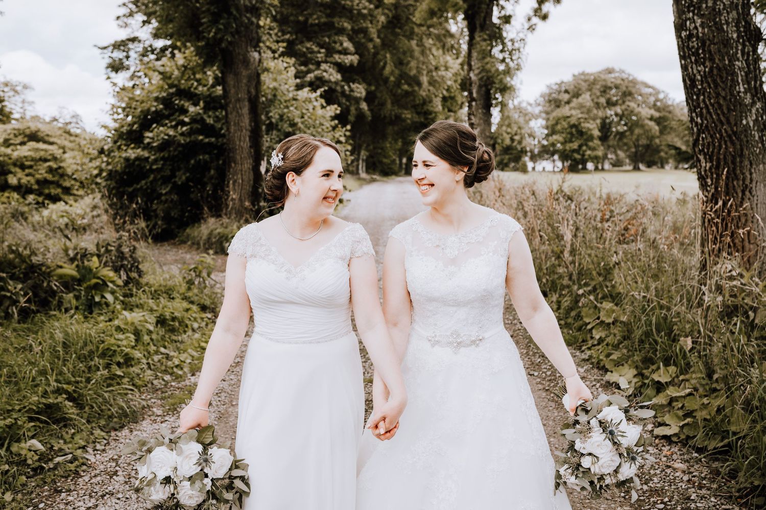 Two people in white wedding dresses walking hand in hand on an outdoor path.