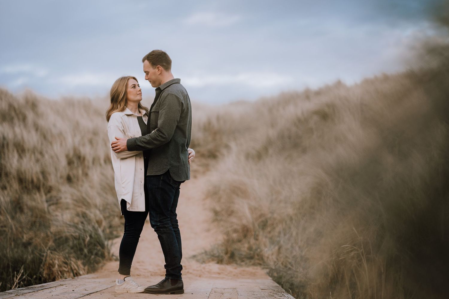A romantic couple embraces on a scenic path surrounded by tall dried grasses on a moody afternoon.